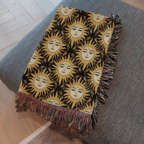 A woven cotton throw featuring a celestial sun face pattern in gold and black tones, framed by soft fringe.